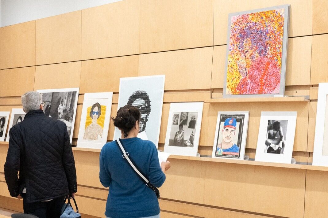 Two people stand and examine framed portraits and colorful artwork displayed on a wooden gallery wall.