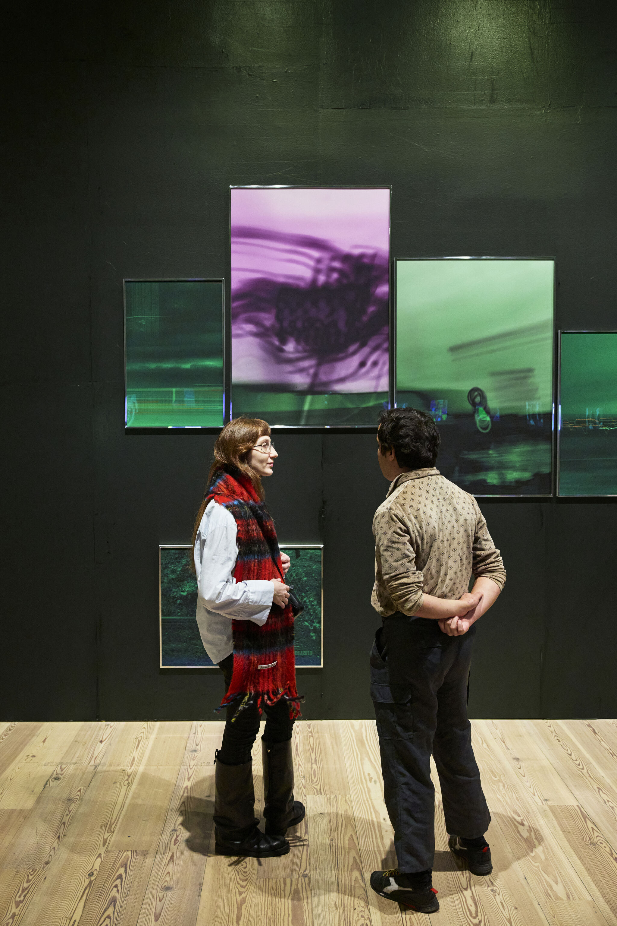 Two people converse in front of a dark wall with a series of abstract photographs with thin chrome frames, comprising glowing green and purple flowing lines.