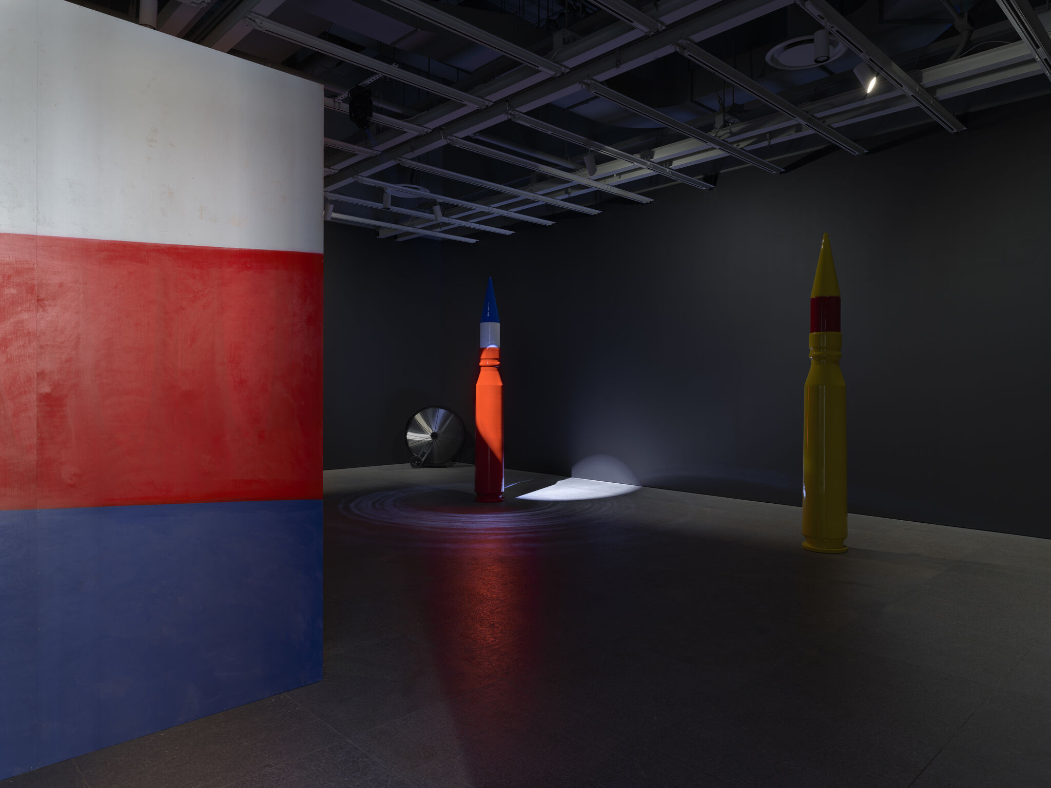 Two tall pencil-shaped sculptures are lit in a dark gallery beside a red and blue wall.