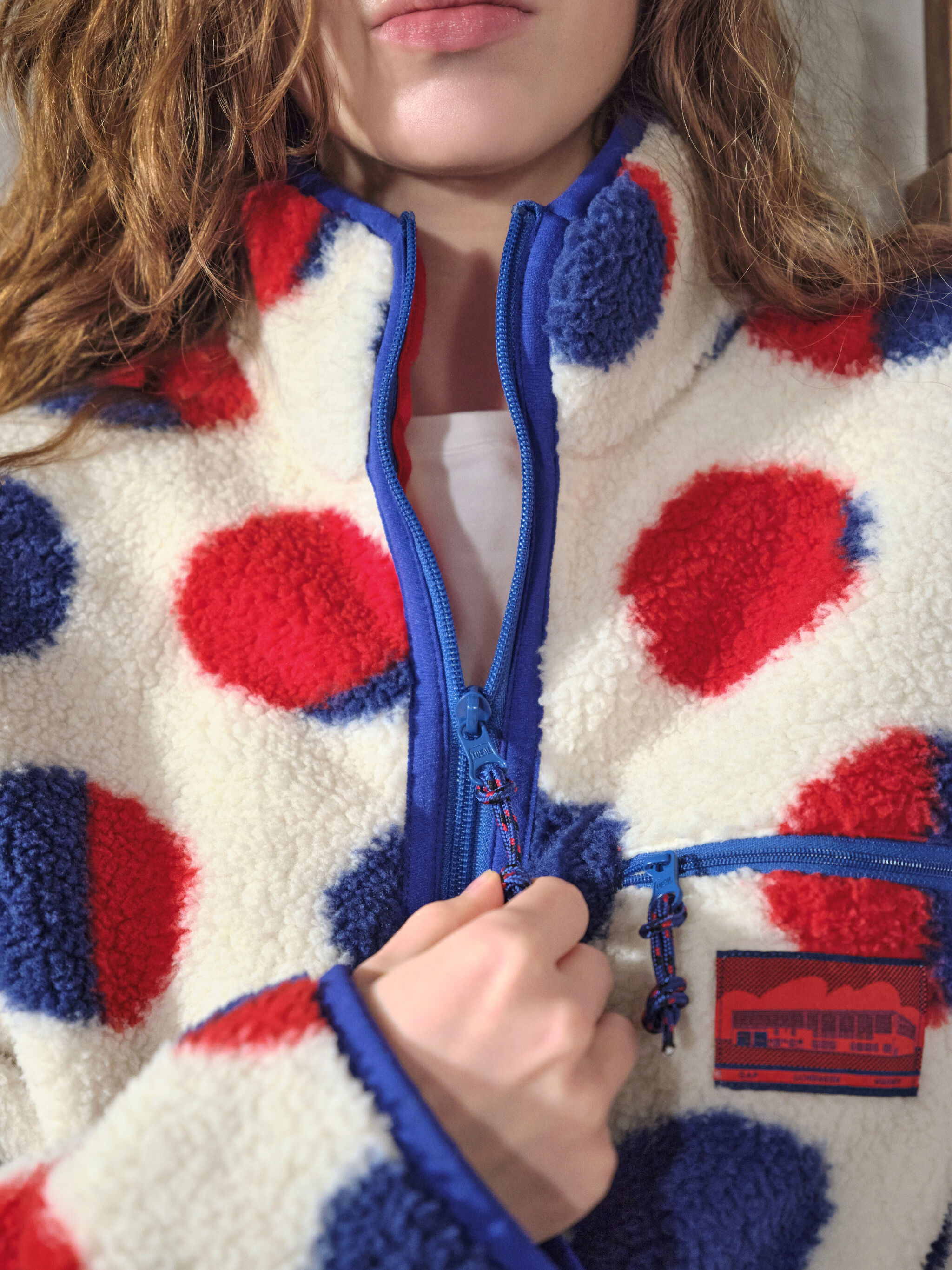 Model zipping a red-and-blue dotted sherpa fleece inspired by Lichtenstein’s Cathedral #2