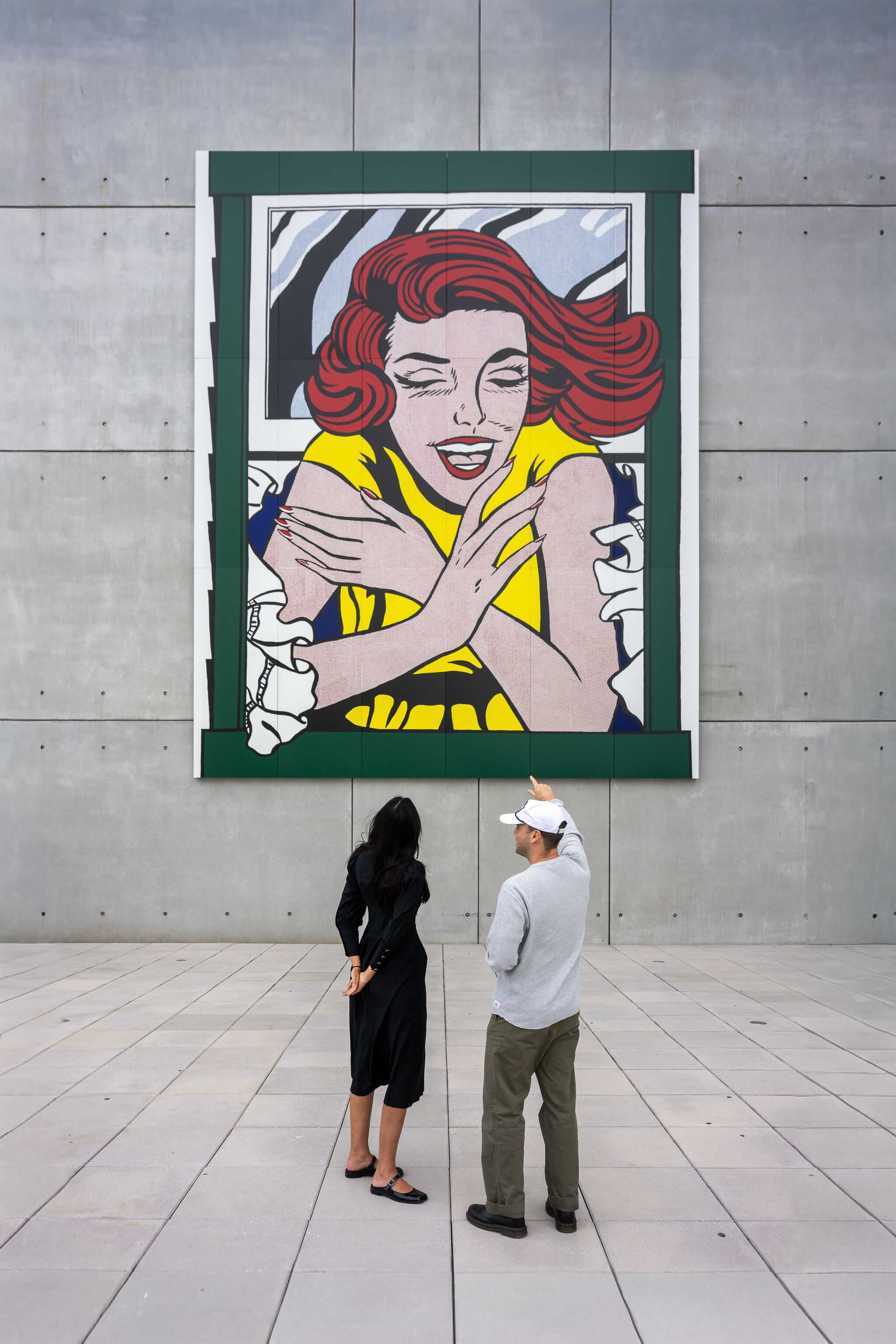 Two people look up at a large pop-art mural of a red-haired woman hugging herself.