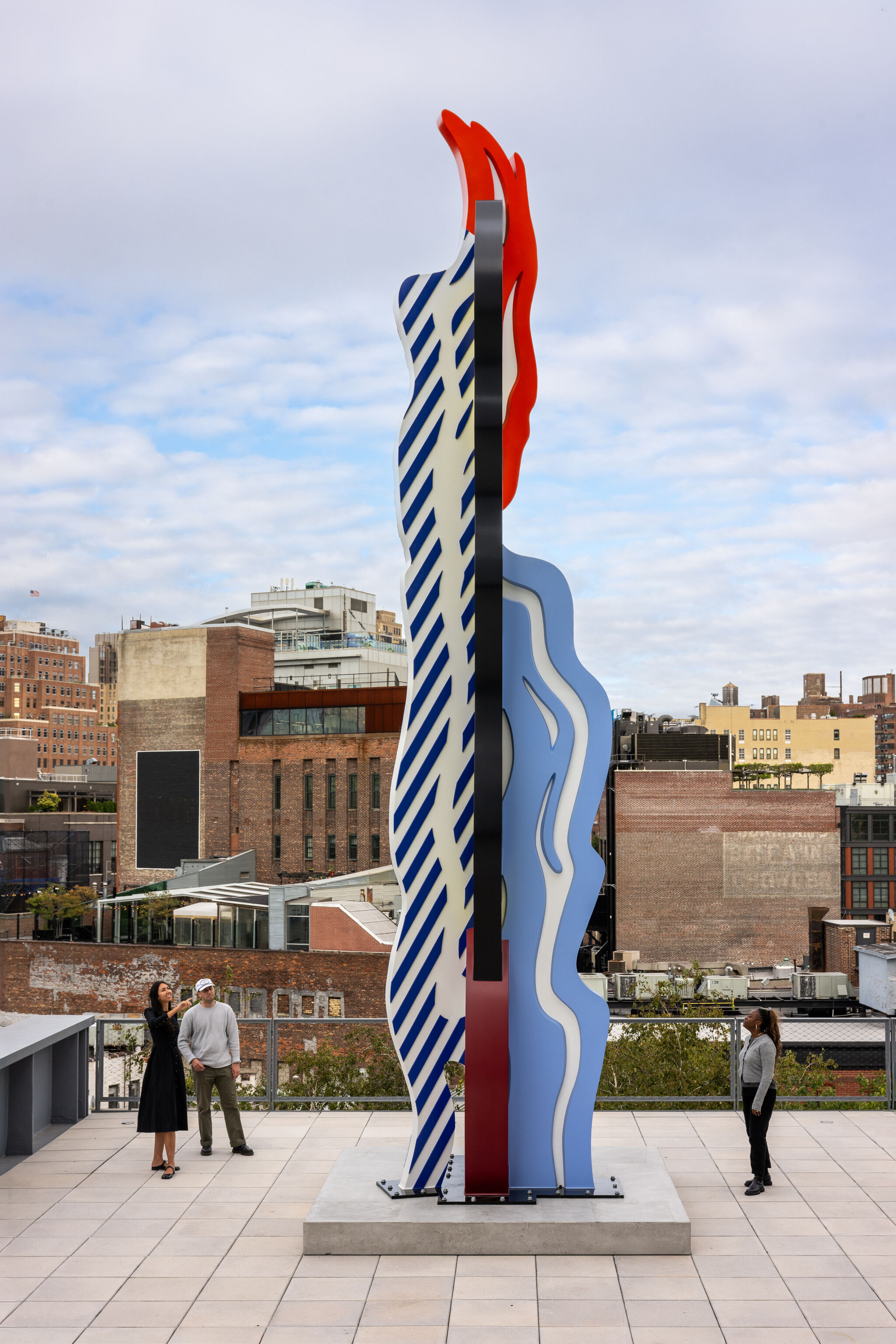 Tall colorful abstract sculpture stands on a rooftop plaza while visitors look and point.