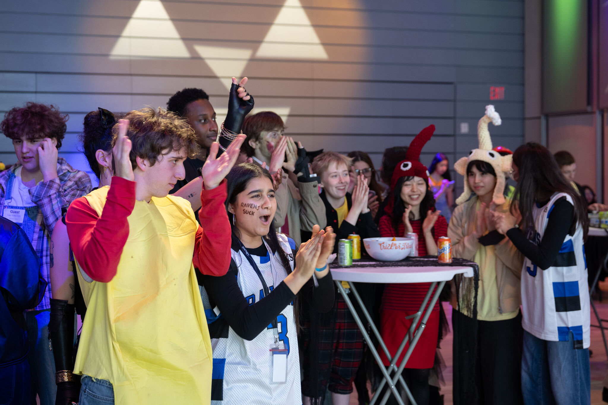 A group of young people in costumes cheer and clap together at an indoor event.