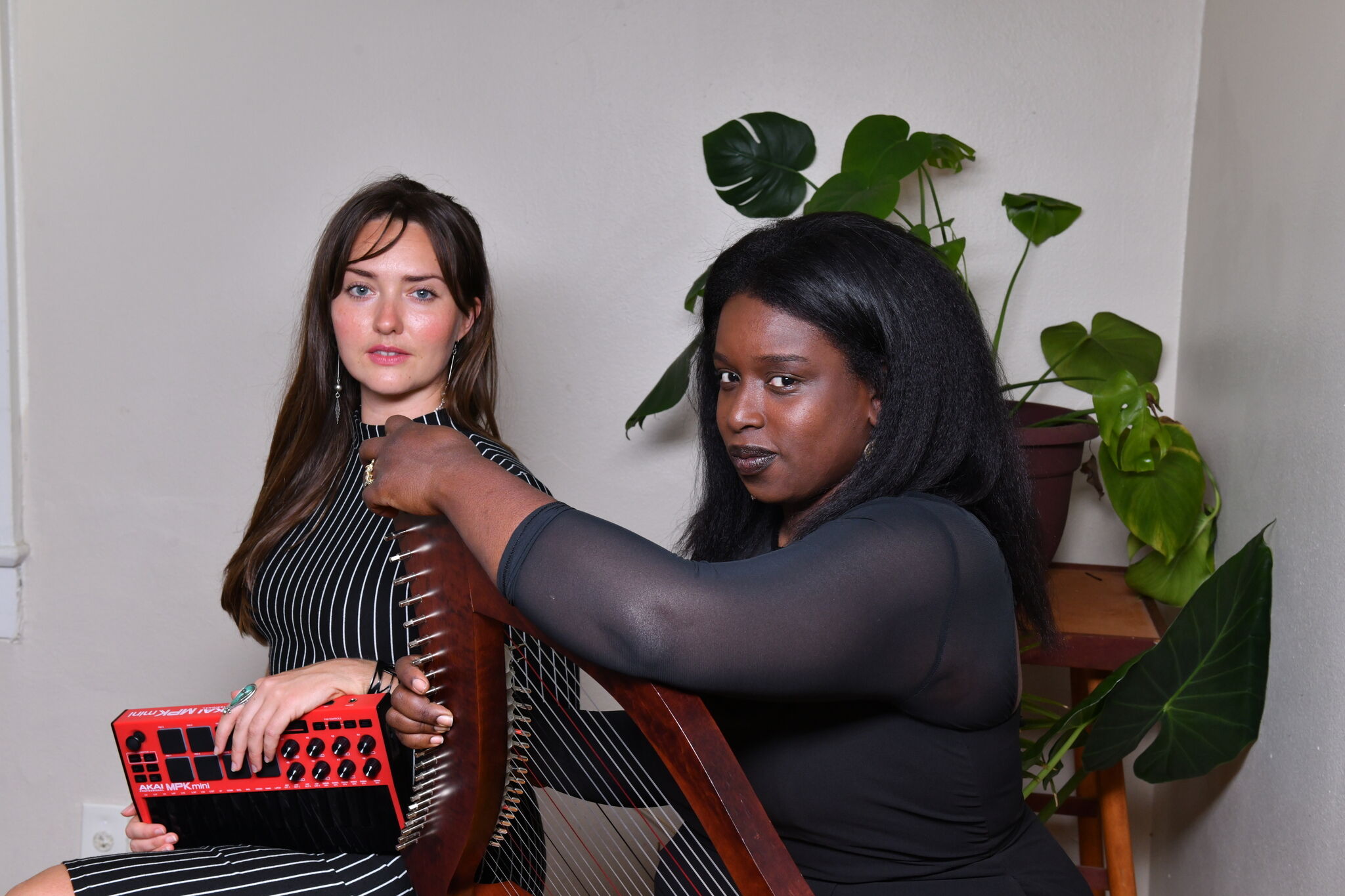 Two women pose with a wooden harp while one holds a red midi controller and plucks strings.