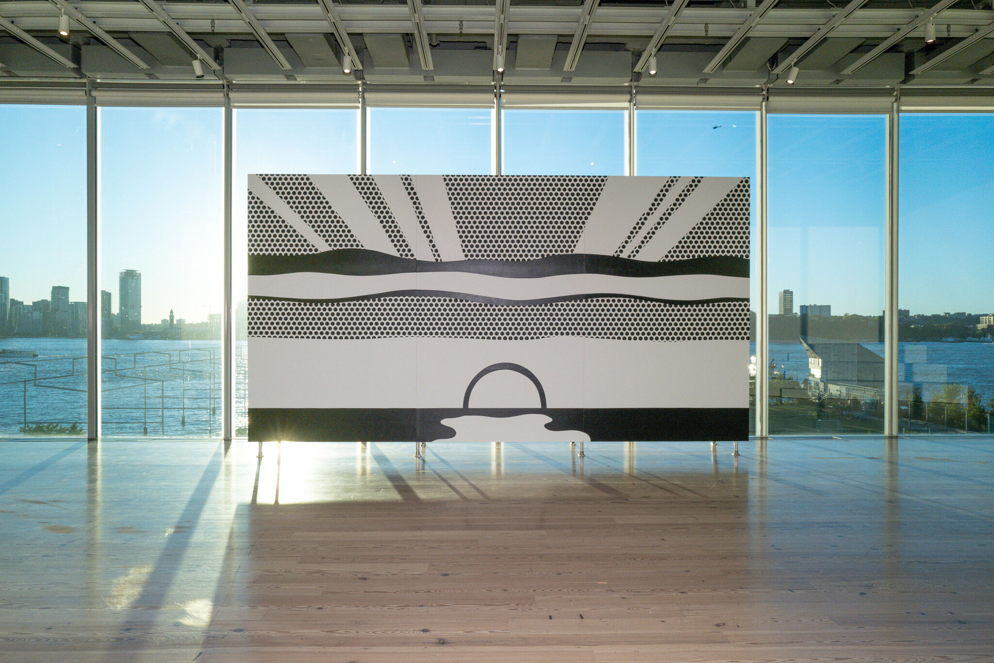 A large black-and-white reproduction of a Roy Lichtenstein landscape is installed in a sunlit gallery at the Whitney Museum. The image features bold horizontal bands, Ben-Day dots, radiating lines, and a simplified setting sun, with floor-to-ceiling windows and the Hudson River visible behind it.