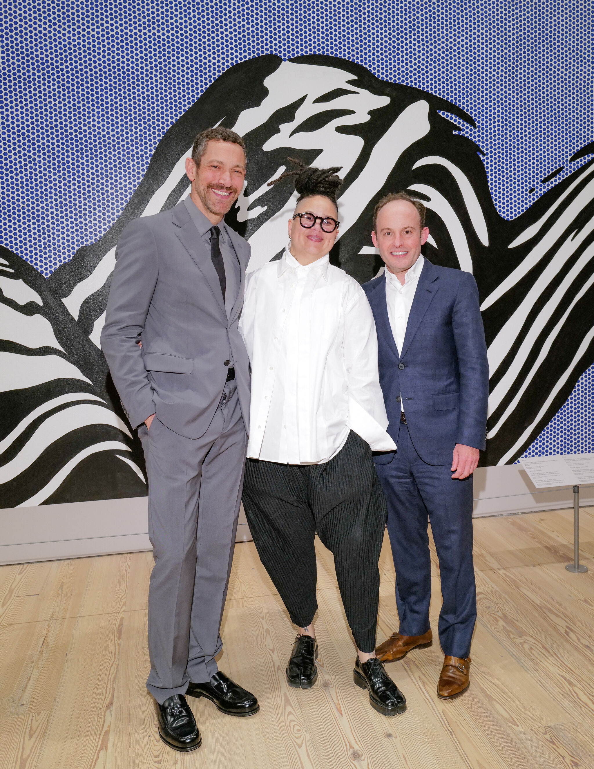 Three people stand smiling together in front of a large black-and-white patterned mural.