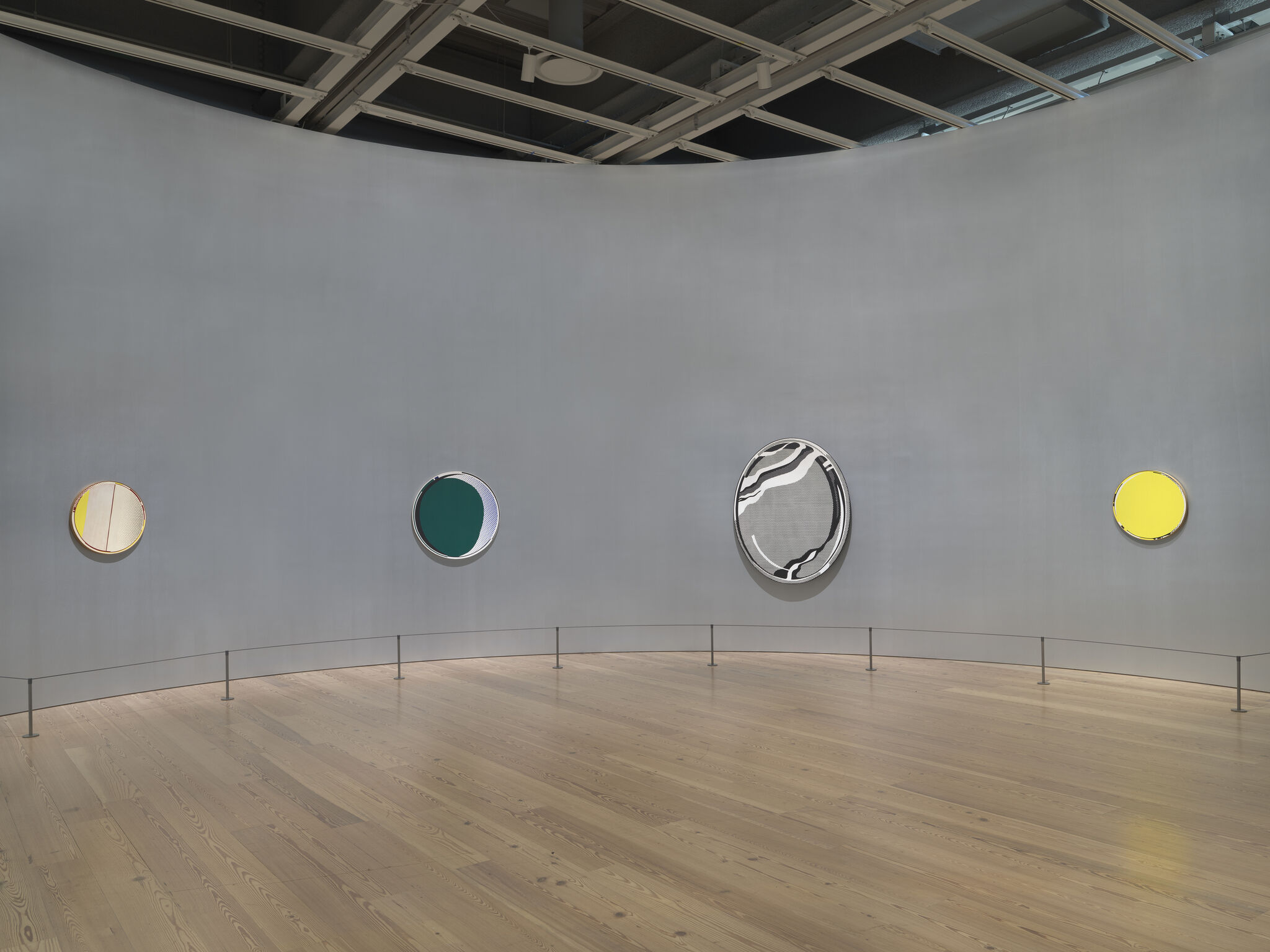 Five round colorful wall artworks mounted on a large curved gray gallery wall above a wooden floor.