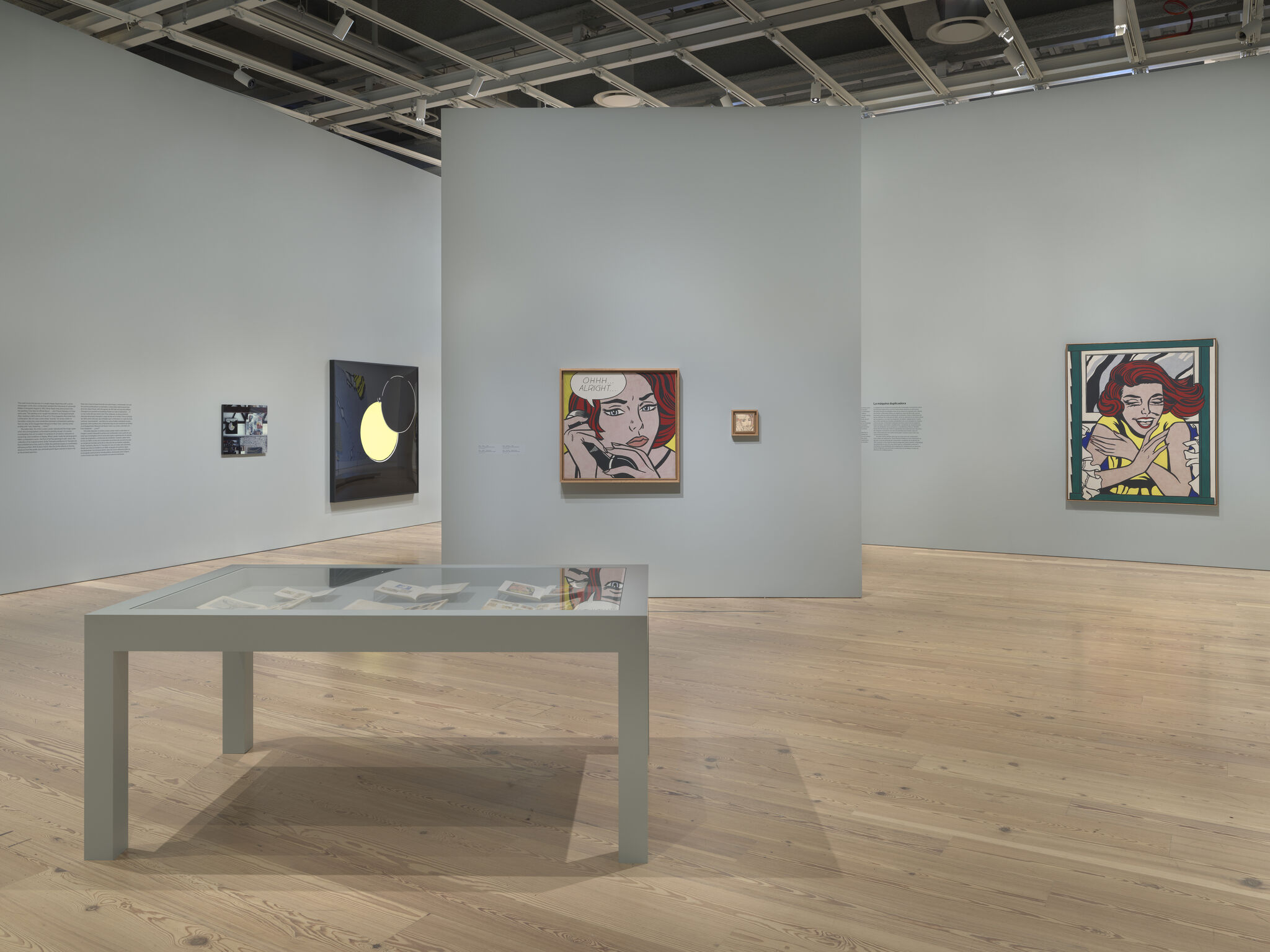 Gallery room with Pop Art paintings on pale walls and a glass display table in foreground.