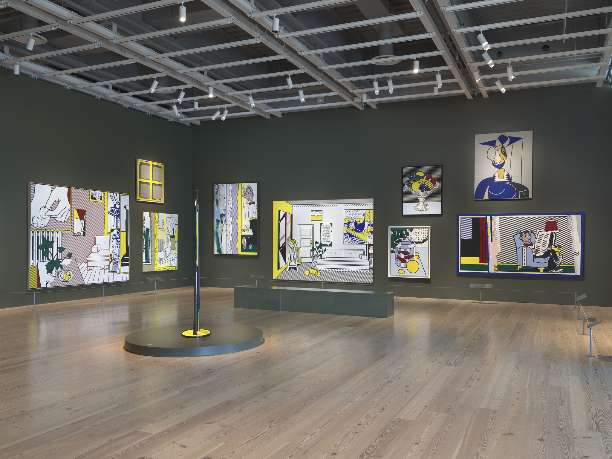 A gallery room display of colorful pop-art paintings on a dark green wall with central sculpture.