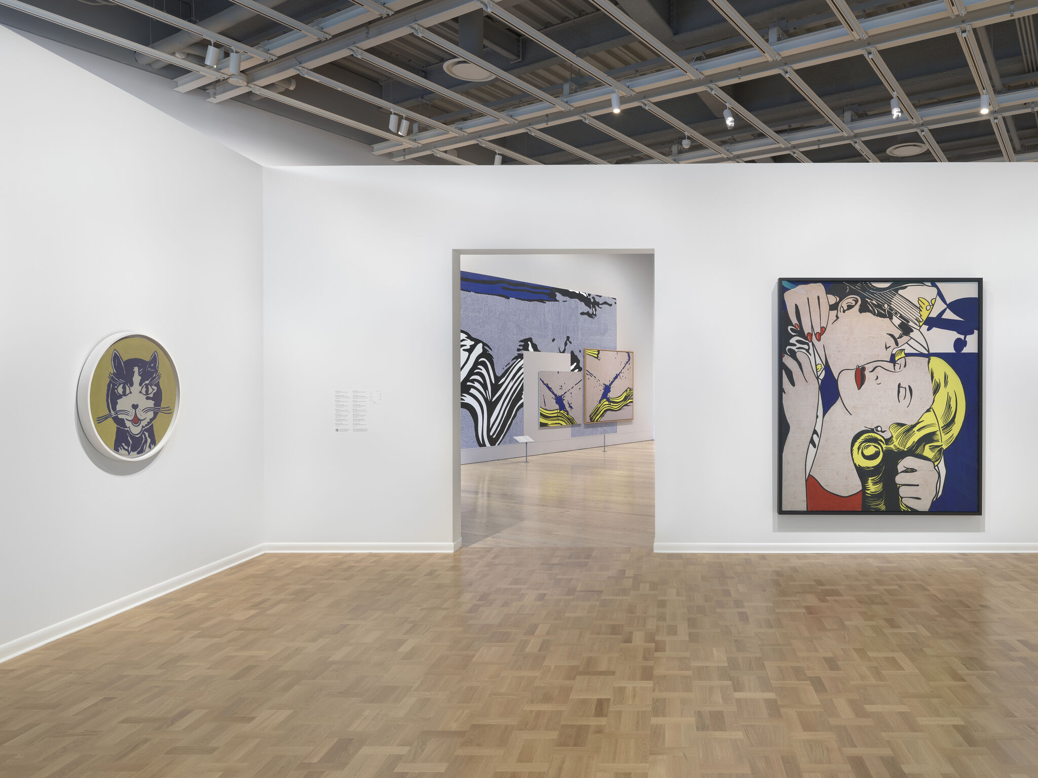 White gallery room showing pop-art paintings, including a large kissing couple and a round cat portrait.
