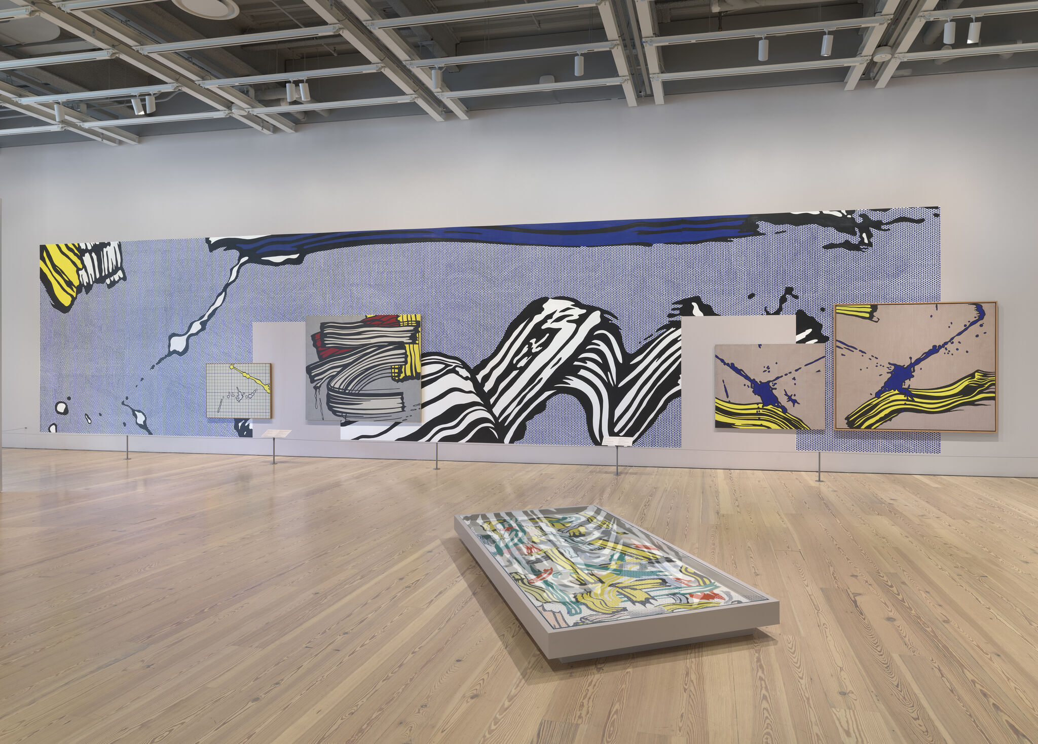 Large colorful pop art mural and framed paintings displayed on a white gallery wall with a floor case.