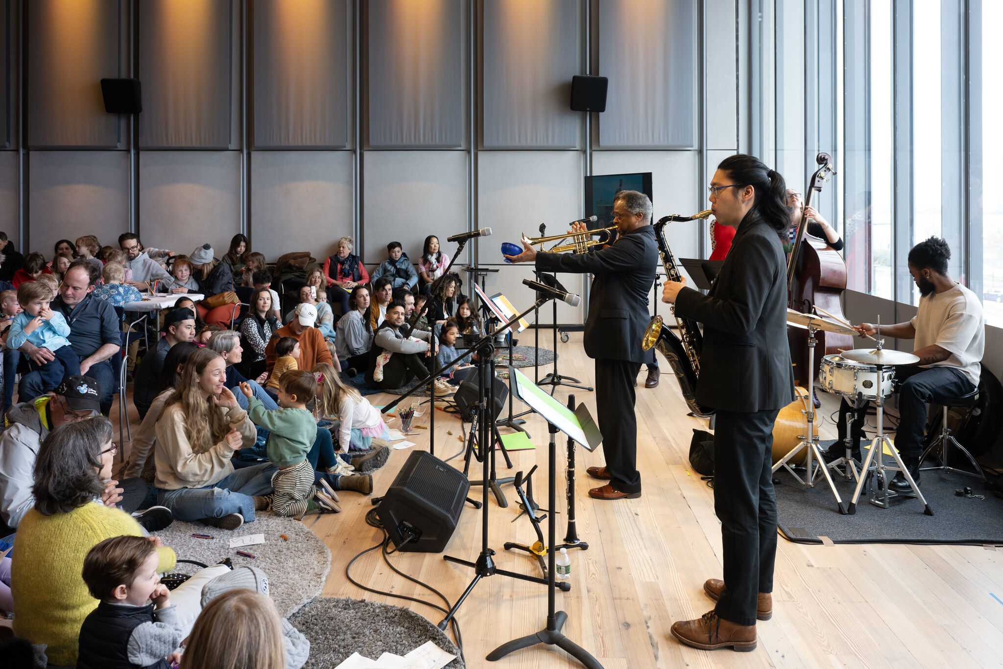 A jazz quartet plays trumpet and saxophone while families and children sit on the floor watching.