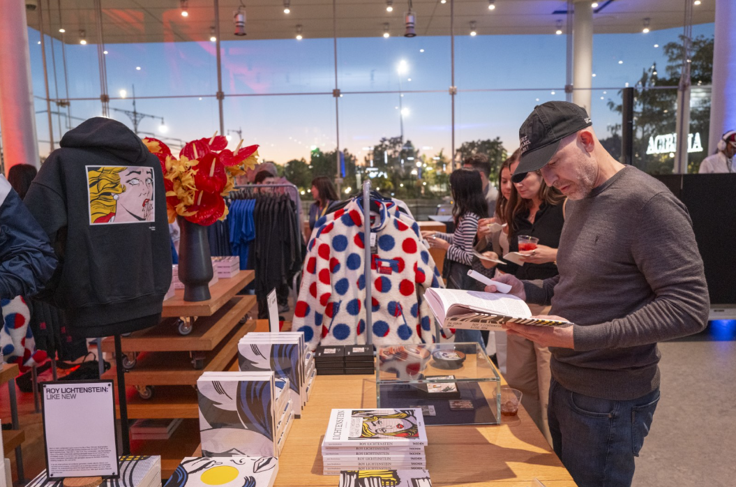 A man in a gray shirt and a gray baseball cap is looking at a book in the Whitney Shop. Roy Lichtenstein merchandise is on display. 