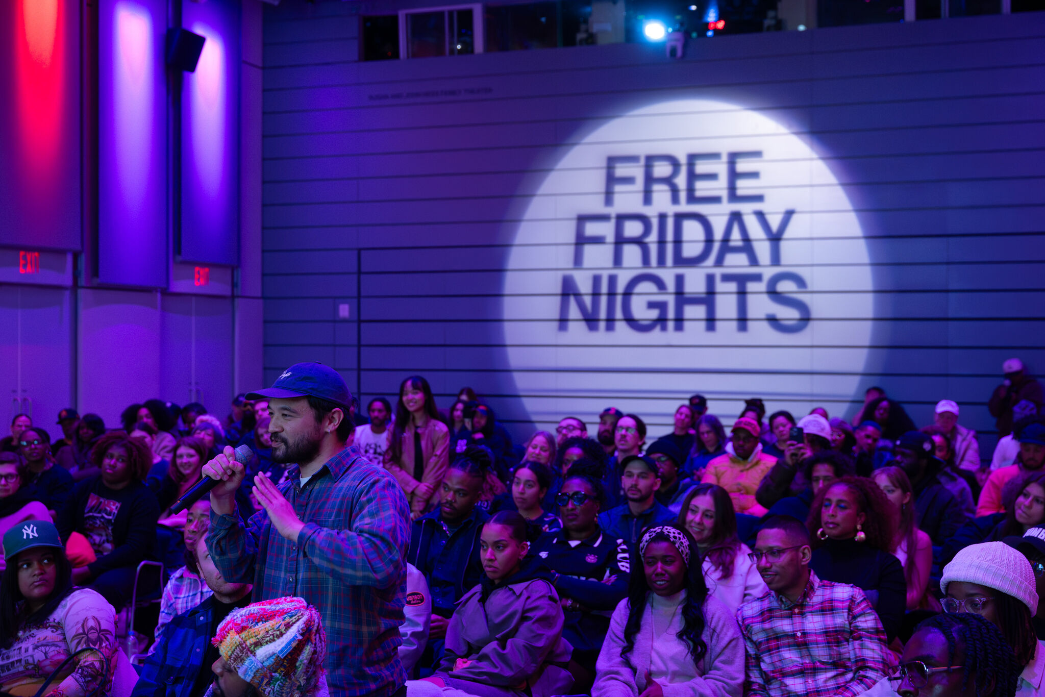 A man speaks into a microphone while a crowd watches and "Free Friday Nights" is projected.